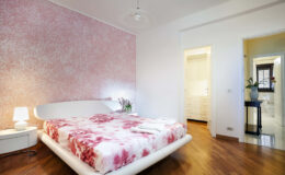 Modern bedroom featuring a stylish white bed with a pink floral bedspread, soft lighting from a bedside lamp, and a textured pink wall. The room includes a doorway leading to an en-suite bathroom and a closet area, with polished wooden flooring enhancing the contemporary design.