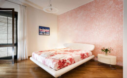 Modern bedroom featuring a floral-patterned bedspread, sleek white bed frame, wooden flooring, and soft pink accent wall. Natural light streams in through large windows with sheer curtains, creating a warm and inviting atmosphere. A small nightstand with a vase of flowers adds a touch of elegance.