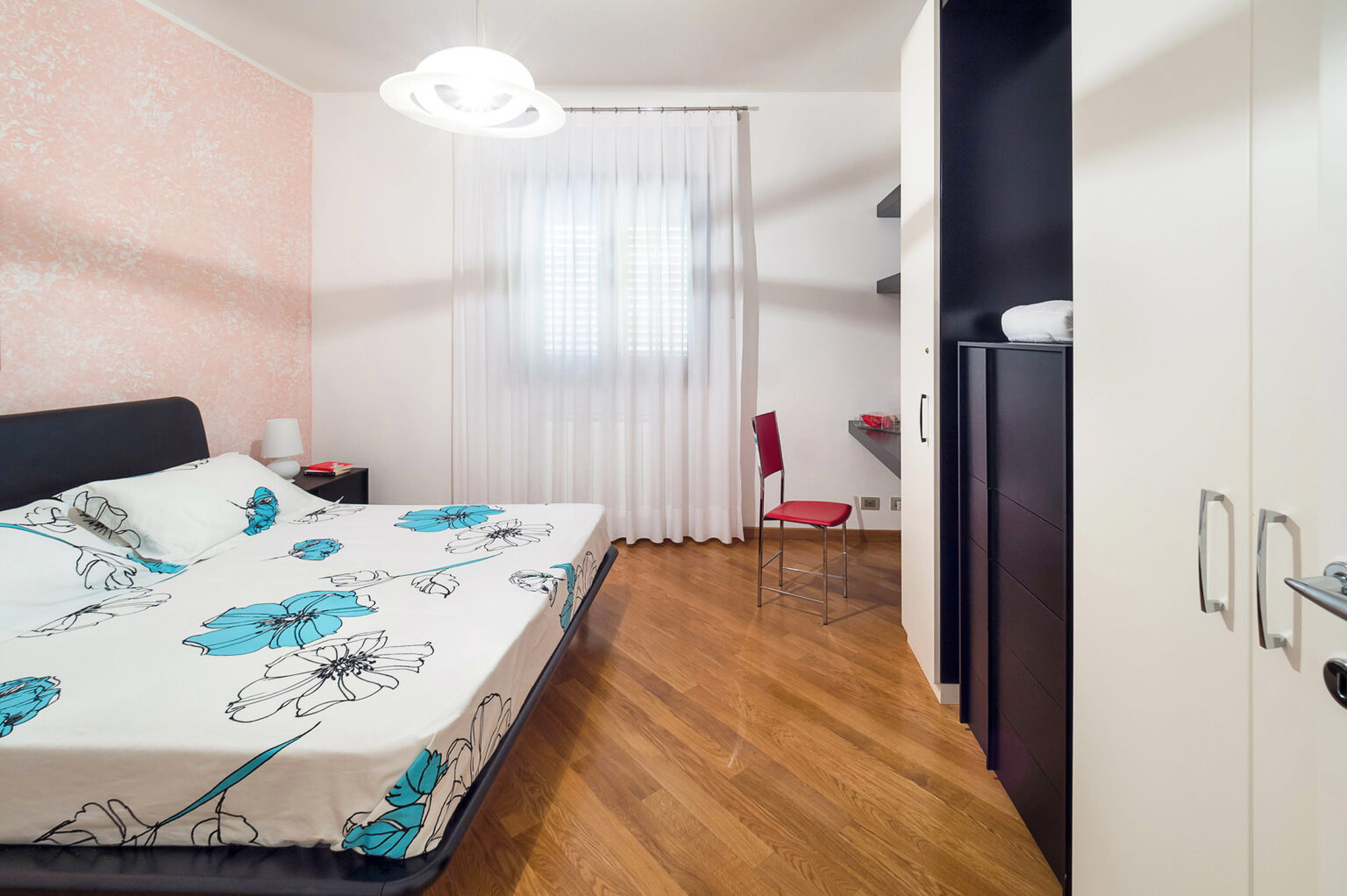 Cozy bedroom featuring a double bed with floral patterned bedding, a modern hanging light fixture, and a red chair. The room has white curtains, a wooden floor, and minimalist furniture, including a nightstand and wardrobe, creating a bright and inviting atmosphere. | Only on SopranoVillas