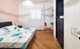Cozy bedroom featuring a double bed with floral patterned bedding, a modern hanging light fixture, and a red chair. The room has white curtains, a wooden floor, and minimalist furniture, including a nightstand and wardrobe, creating a bright and inviting atmosphere.