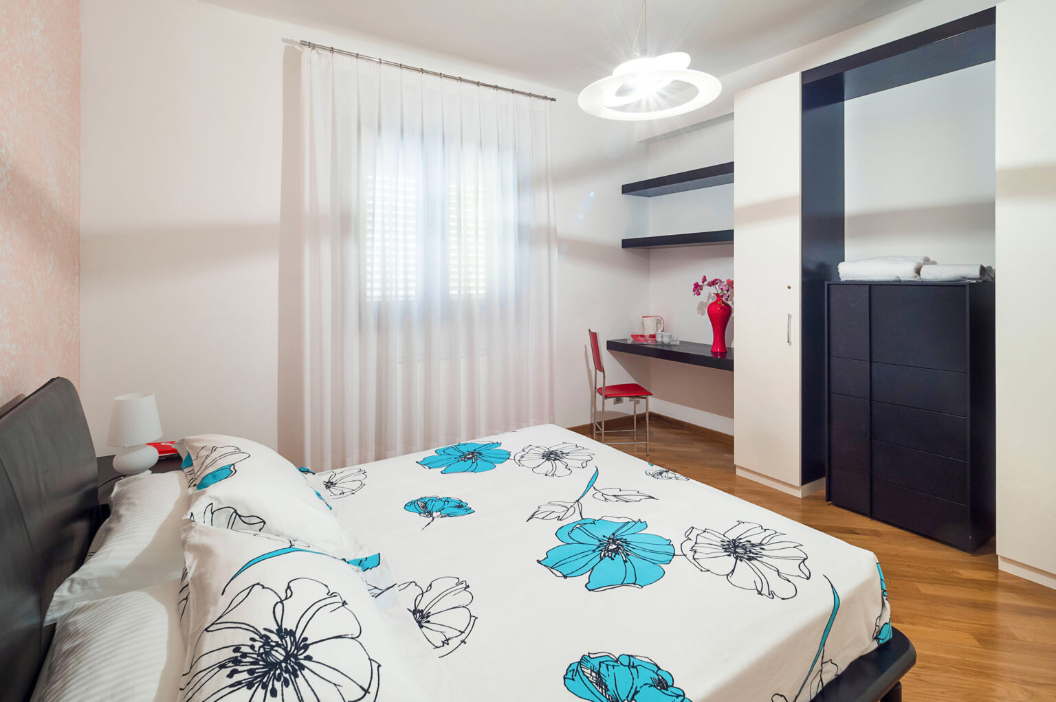 Cozy modern bedroom featuring a floral patterned bedspread, wooden flooring, and a stylish chandelier. The room includes a desk area with a chair and shelves, complemented by natural light from a large window with sheer curtains. | Only on SopranoVillas