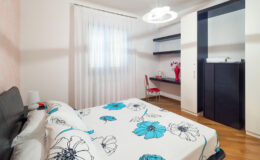 Cozy modern bedroom featuring a floral patterned bedspread, wooden flooring, and a stylish chandelier. The room includes a desk area with a chair and shelves, complemented by natural light from a large window with sheer curtains.