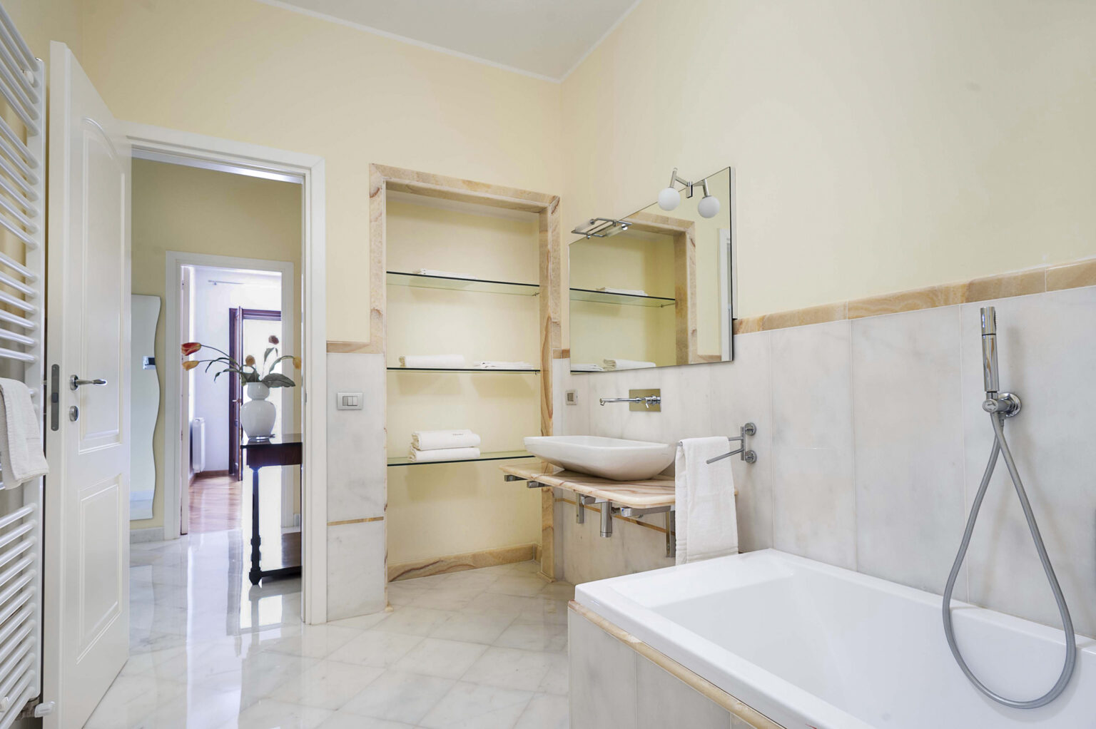 Modern bathroom interior featuring a white bathtub, sleek sink, and minimalist shelving. The walls are painted a soft yellow, complemented by marble flooring. Natural light streams in from an adjacent room, enhancing the spacious feel. | Only on SopranoVillas