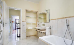 Modern bathroom interior featuring a white bathtub, sleek sink, and minimalist shelving. The walls are painted a soft yellow, complemented by marble flooring. Natural light streams in from an adjacent room, enhancing the spacious feel.