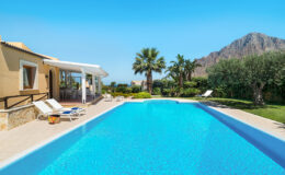 Luxurious villa with a sparkling blue swimming pool, surrounded by palm trees and lush greenery, set against a backdrop of mountains under a clear blue sky. Perfect for a relaxing getaway.