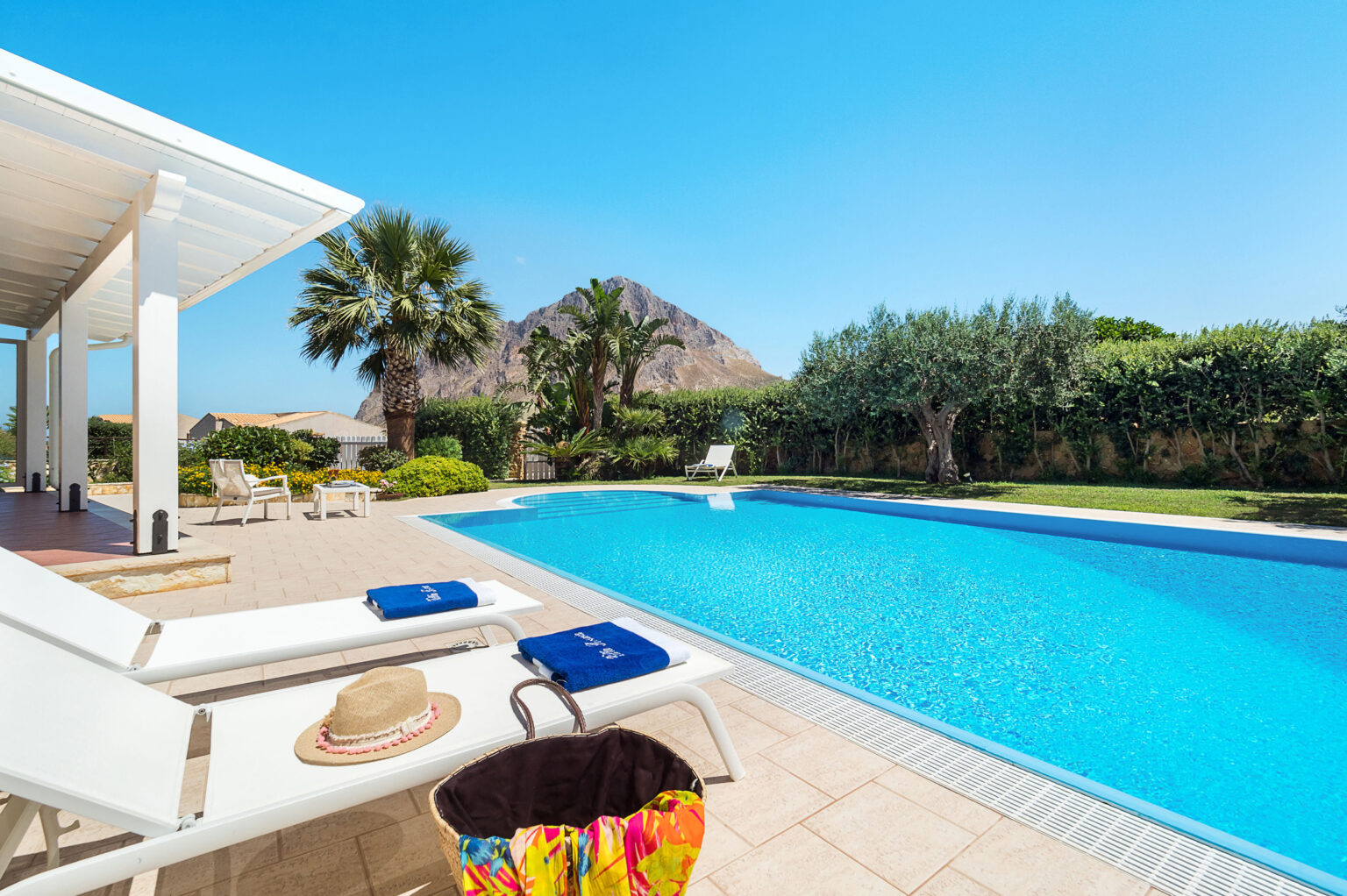 Luxurious outdoor pool area with sun loungers, a straw hat, and a colorful beach bag, surrounded by lush greenery and a mountain backdrop under a clear blue sky. Perfect for relaxation and summer getaways. | Only on SopranoVillas