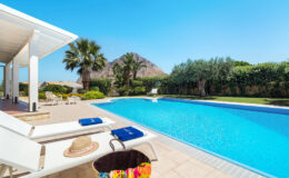 Luxurious outdoor pool area with sun loungers, a straw hat, and a colorful beach bag, surrounded by lush greenery and a mountain backdrop under a clear blue sky. Perfect for relaxation and summer getaways.