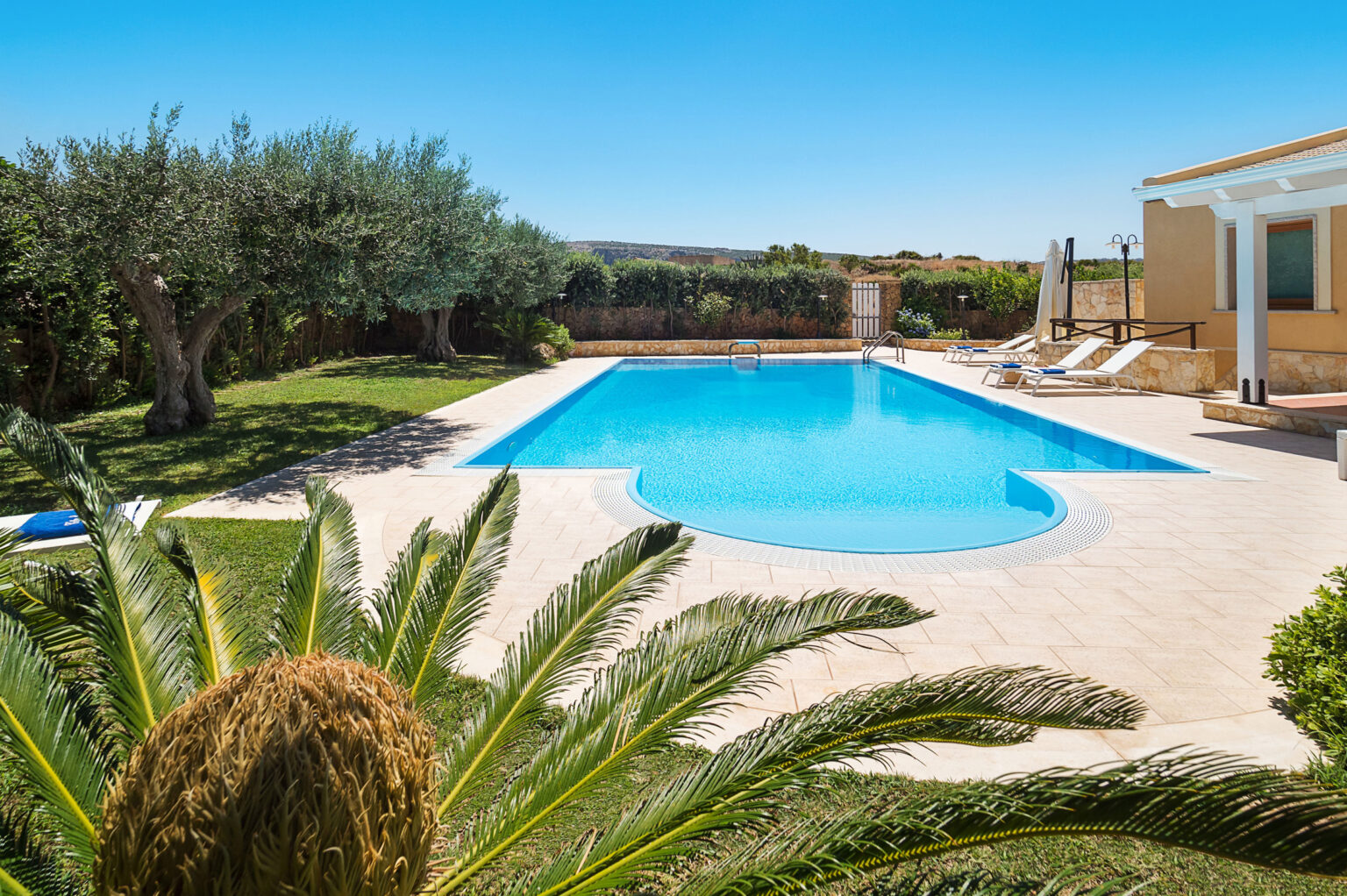 Luxurious outdoor swimming pool surrounded by lush greenery and palm trees, with sun loungers on the patio, ideal for relaxation in a sunny setting. | Only on SopranoVillas