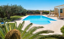 Luxurious outdoor swimming pool surrounded by lush greenery and palm trees, with sun loungers on the patio, ideal for relaxation in a sunny setting.