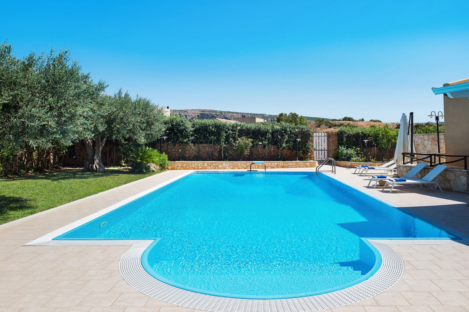 A serene outdoor swimming pool surrounded by lush greenery and olive trees, featuring comfortable lounge chairs and a clear blue sky. Ideal for relaxation and leisure activities. | Only on SopranoVillas