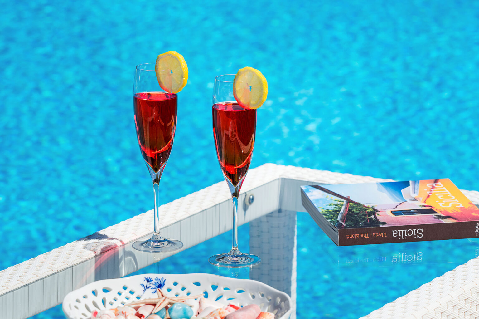Two vibrant red cocktails with lemon slices sit on a table beside a sparkling blue swimming pool, accompanied by a colorful book, creating a relaxing summer vibe. | Only on SopranoVillas