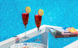 Two vibrant red cocktails with lemon slices sit on a table beside a sparkling blue swimming pool, accompanied by a colorful book, creating a relaxing summer vibe.