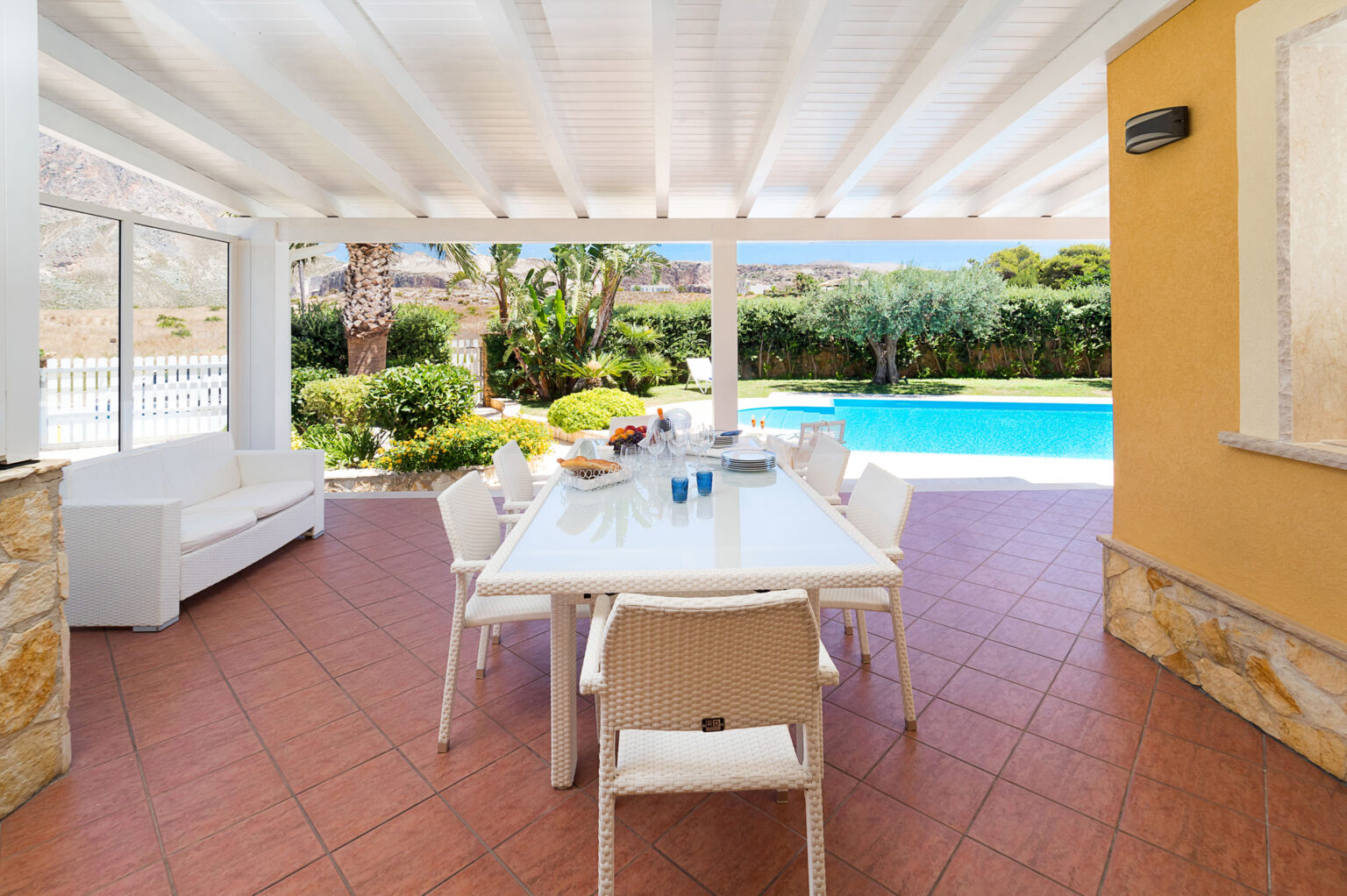 Outdoor dining area with a modern white table and chairs, surrounded by lush greenery and a swimming pool, under a covered patio with a bright, inviting atmosphere. Perfect for relaxation and entertaining. | Only on SopranoVillas