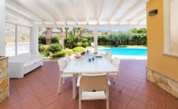 Outdoor dining area with a modern white table and chairs, surrounded by lush greenery and a swimming pool, under a covered patio with a bright, inviting atmosphere. Perfect for relaxation and entertaining.