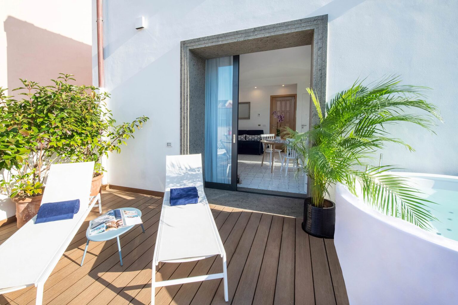 Cozy outdoor patio featuring two white lounge chairs with blue towels, a small coffee table with magazines, and lush greenery, leading to an inviting indoor space with a dining area and a modern hot tub. Perfect for relaxation and leisure. | Only on SopranoVillas