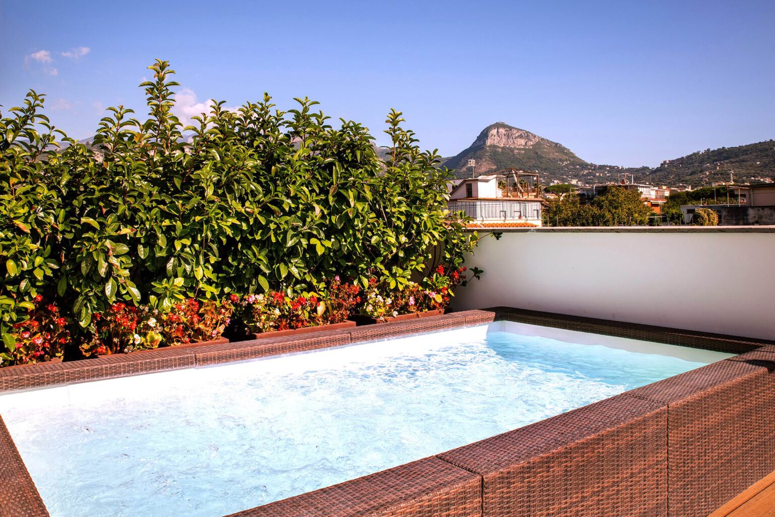 Rooftop pool surrounded by lush greenery and colorful flowers, with a scenic mountain view in the background under a clear blue sky. Ideal for relaxation and outdoor enjoyment. | Only on SopranoVillas
