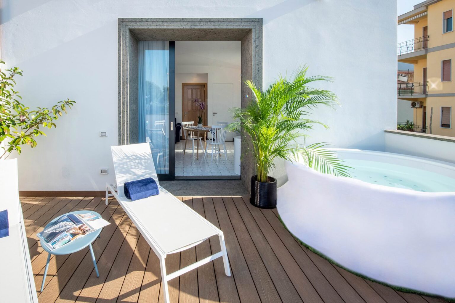Modern outdoor balcony featuring a sun lounger, a small table with a magazine, a lush potted plant, and a round hot tub, with a view into a bright indoor living space. Ideal for relaxation and leisure. | Only on SopranoVillas