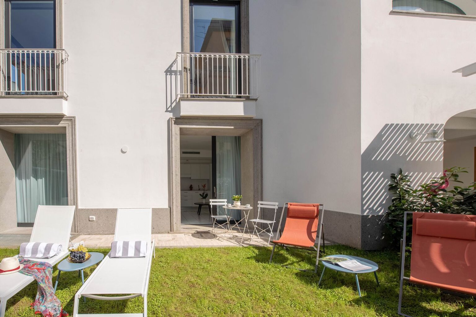 Modern outdoor patio area with lounge chairs and a small table, featuring a doorway leading into a bright kitchen. Lush green grass and decorative plants enhance the tranquil ambiance. Ideal for relaxation and outdoor dining. | Only on SopranoVillas