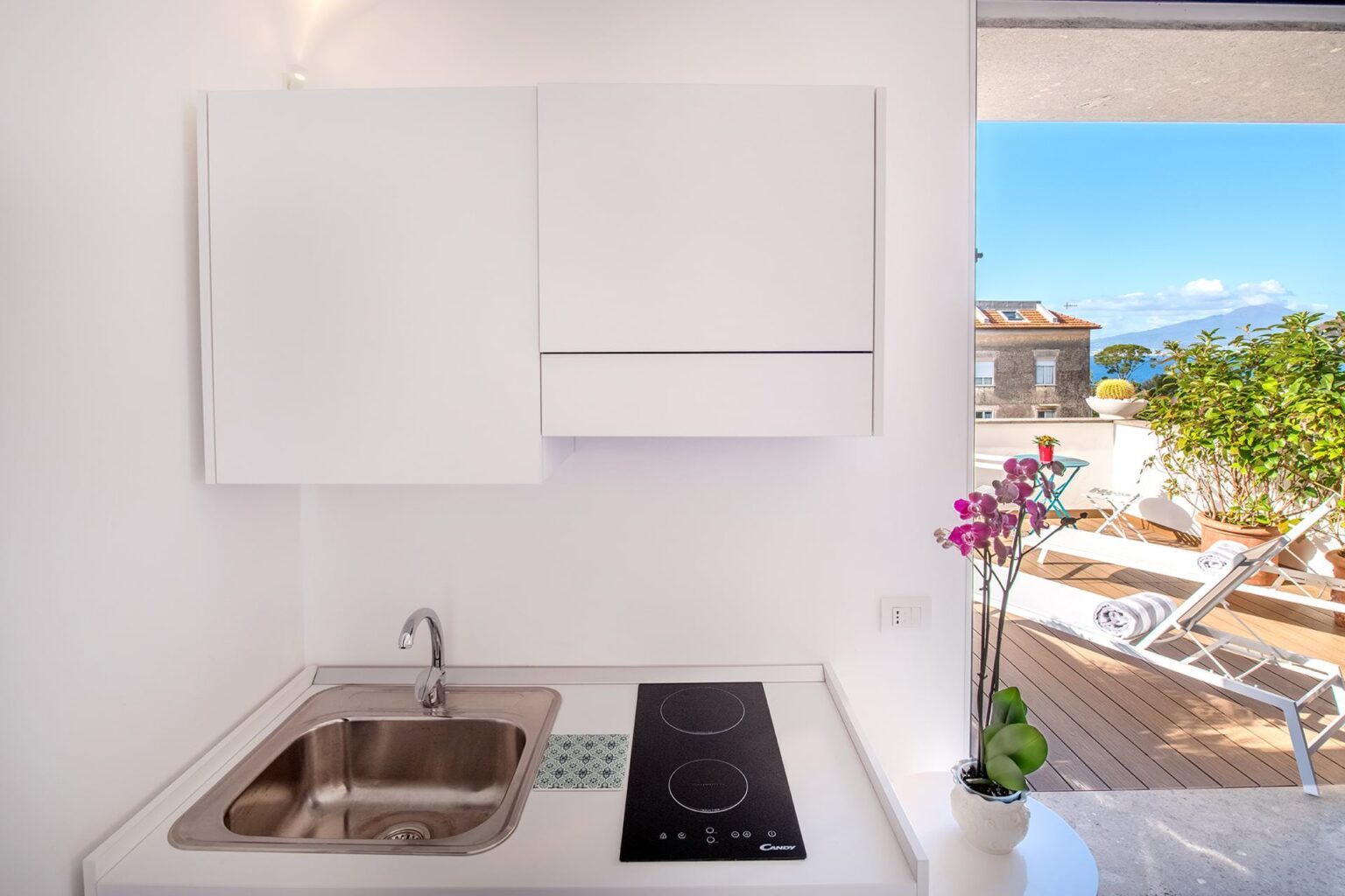 Modern kitchen interior featuring a stainless steel sink, electric cooktop, and white cabinetry. A scenic view of outdoor seating and greenery is visible through the window, enhancing the bright and airy atmosphere. | Only on SopranoVillas