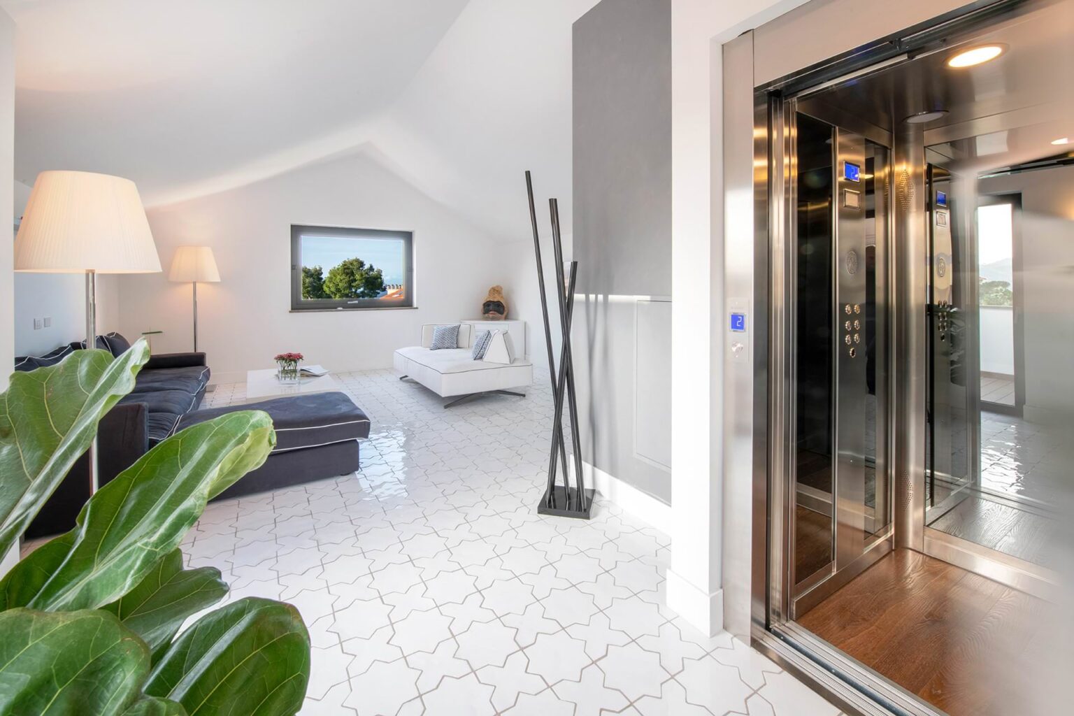 Modern interior of a bright living space featuring an elevator, stylish furniture, and large windows with a scenic view. The room has a contemporary design with white tiles and decorative plants. | Only on SopranoVillas