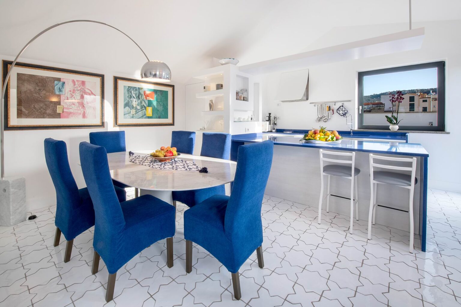 Modern dining area featuring a round table surrounded by six blue upholstered chairs, complemented by colorful artwork on the walls. The open kitchen is visible in the background, showcasing a sleek design with a blue countertop and bar seating. Natural light floods the space through a large window, enhancing the bright and inviting atmosphere. | Only on SopranoVillas