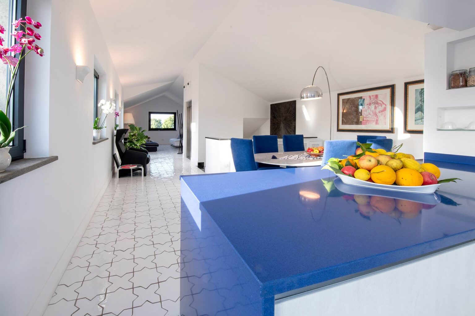 Bright and modern kitchen featuring a blue countertop, white walls, and decorative fruit bowl. The open layout includes a dining area with blue chairs and framed artwork, complemented by natural light from large windows. | Only on SopranoVillas