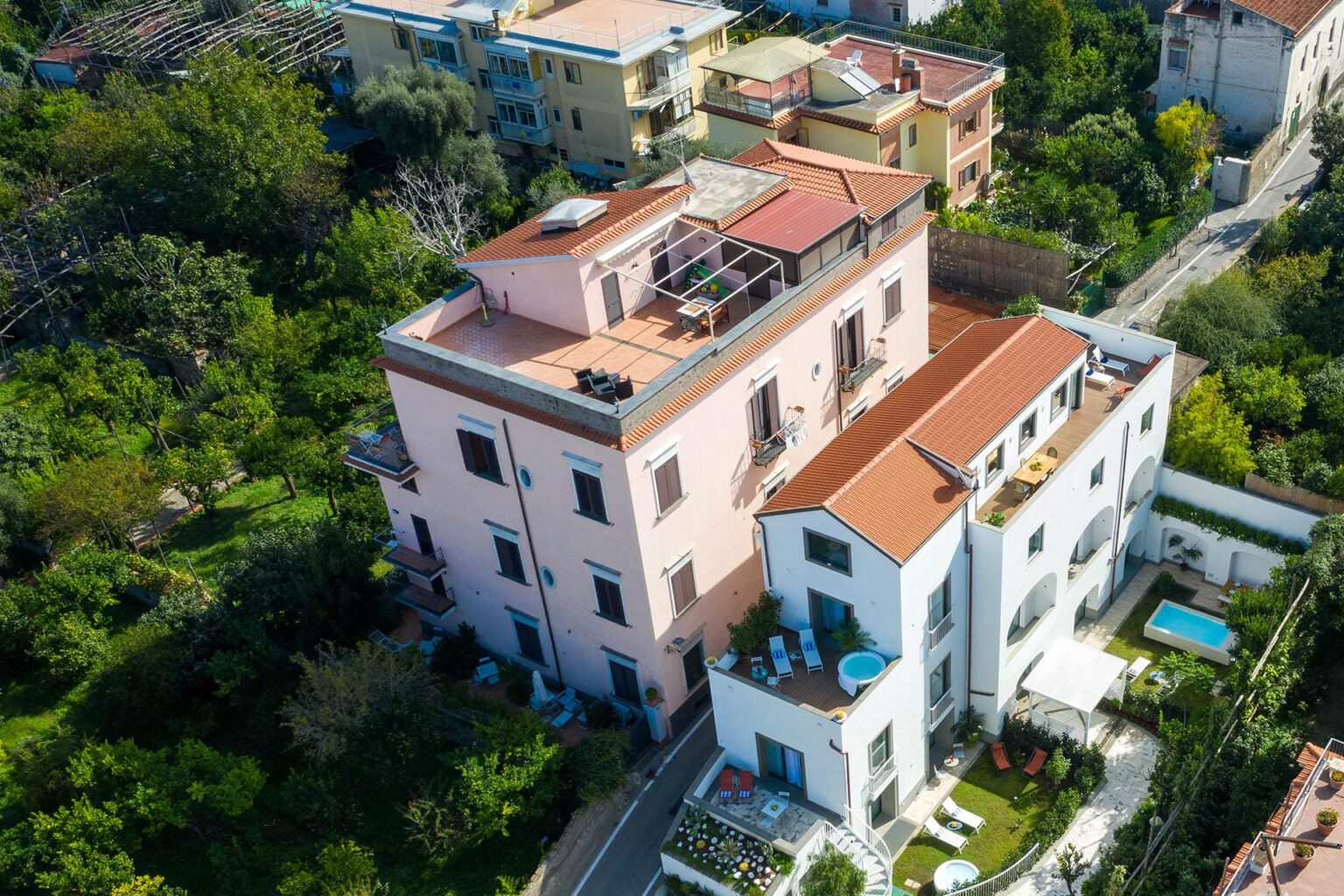 Aerial view of a modern multi-story building with colorful facades surrounded by lush greenery, showcasing a rooftop terrace and swimming pool in a residential area. | Only on SopranoVillas