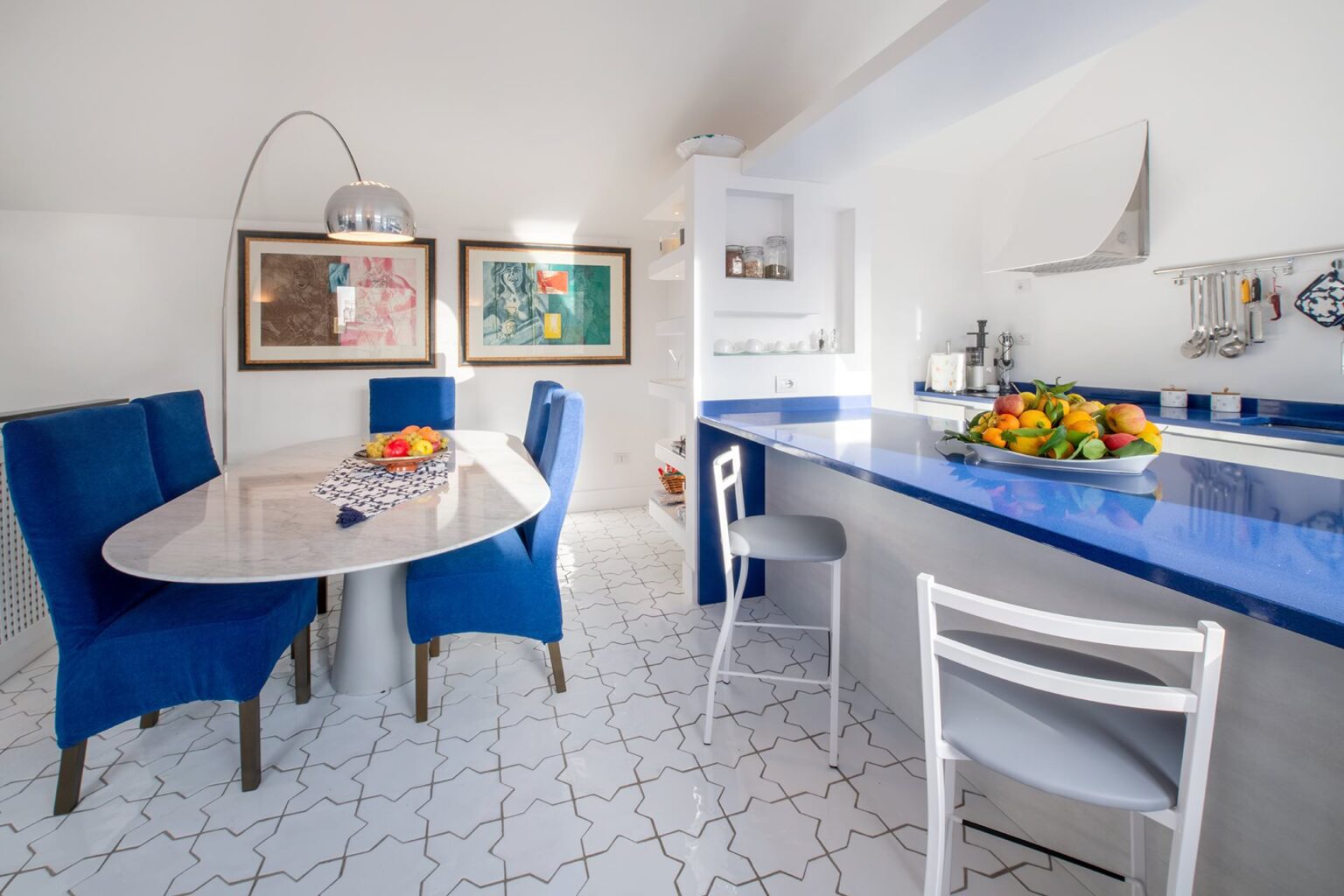 Modern kitchen interior featuring a bright blue countertop, a round dining table with blue chairs, and white tile flooring. The space is decorated with colorful artwork and a fruit bowl, creating a vibrant and inviting atmosphere. | Only on SopranoVillas