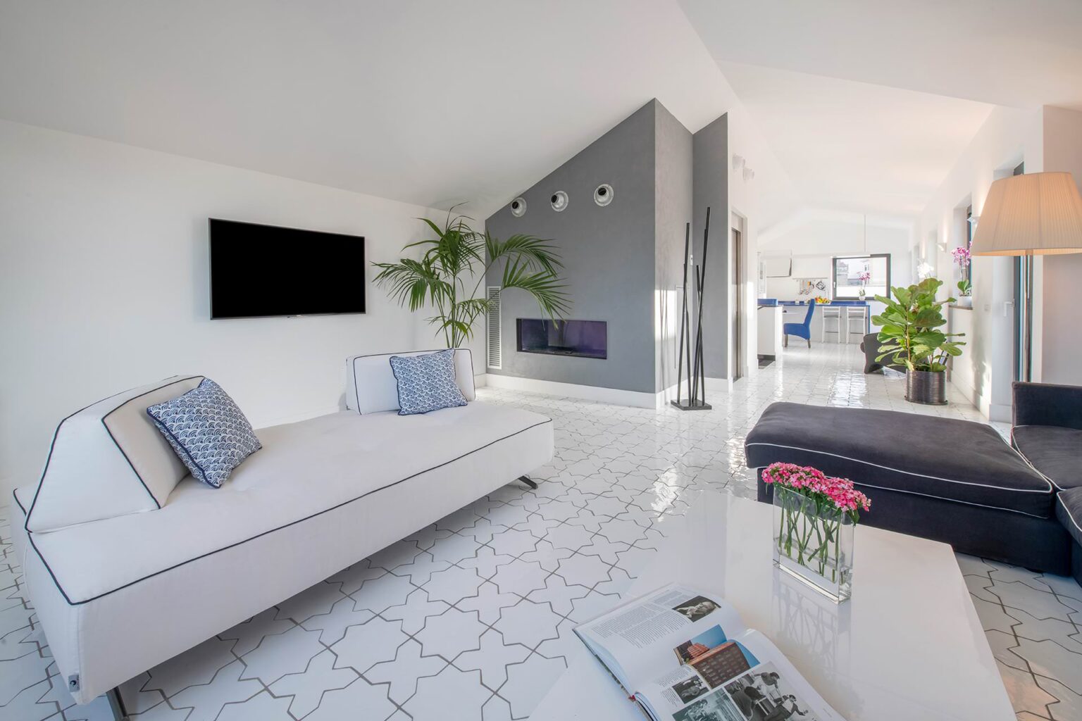 Modern living room featuring a white sofa, a sleek fireplace, and a large flat-screen TV, complemented by indoor plants and stylish decor. Bright, open space with elegant tile flooring and contemporary furnishings. | Only on SopranoVillas