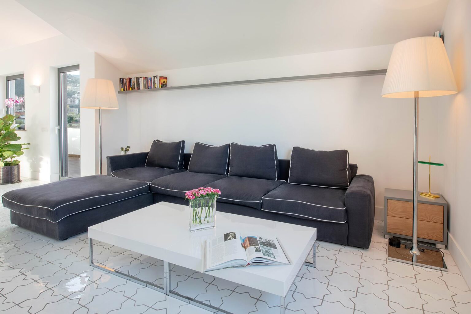 Modern living room featuring a dark gray sectional sofa, a white coffee table with a magazine, and stylish floor lamps. The space is bright with natural light and has minimalist decor, creating a welcoming atmosphere. | Only on SopranoVillas