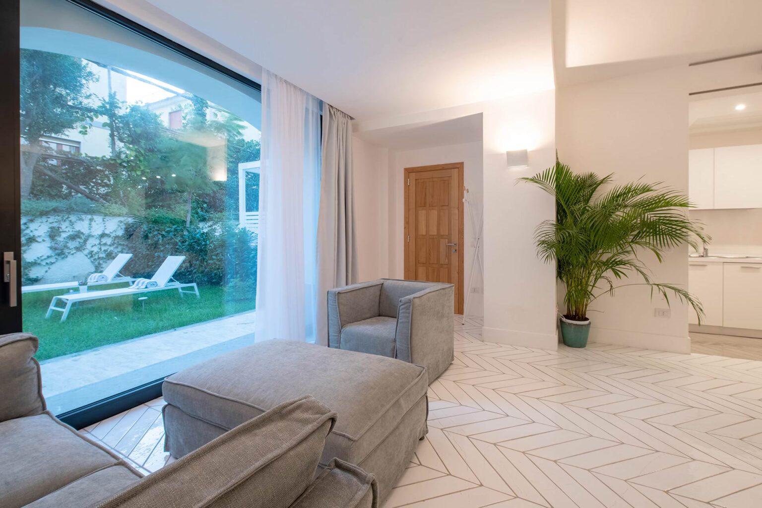 Modern living room with large glass windows overlooking a garden, featuring a comfortable gray sofa, stylish decor, and a potted plant, creating a bright and inviting atmosphere. | Only on SopranoVillas