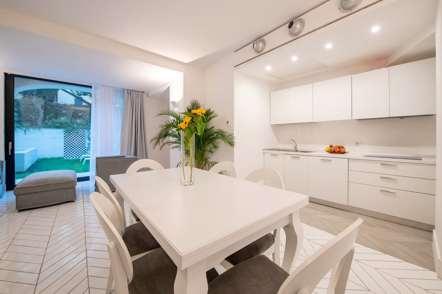 Modern kitchen and dining area featuring a white table, stylish chairs, and a large window with natural light. The kitchen has sleek white cabinets and a fruit bowl on the counter, with a cozy seating area visible through the glass doors. | Only on SopranoVillas