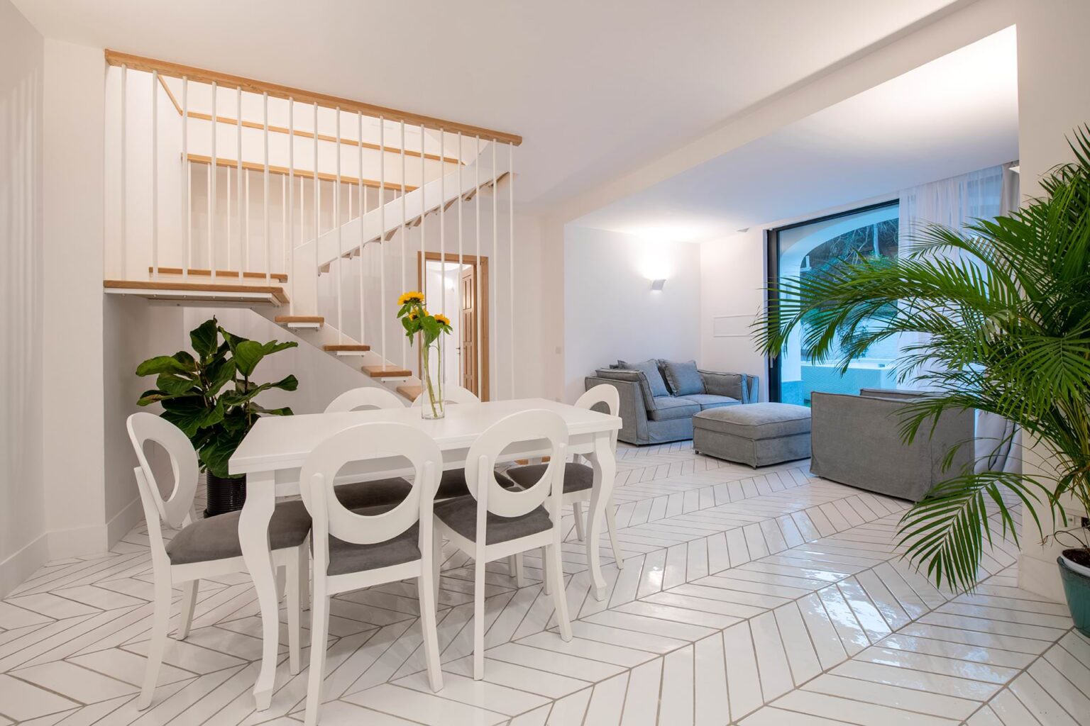 Modern dining area featuring a white table and chairs, surrounded by stylish decor. A staircase leads to the upper level, while a cozy living space with gray sofas is visible in the background. Lush green plants add a touch of nature, and the elegant herringbone-patterned floor enhances the contemporary aesthetic. | Only on SopranoVillas