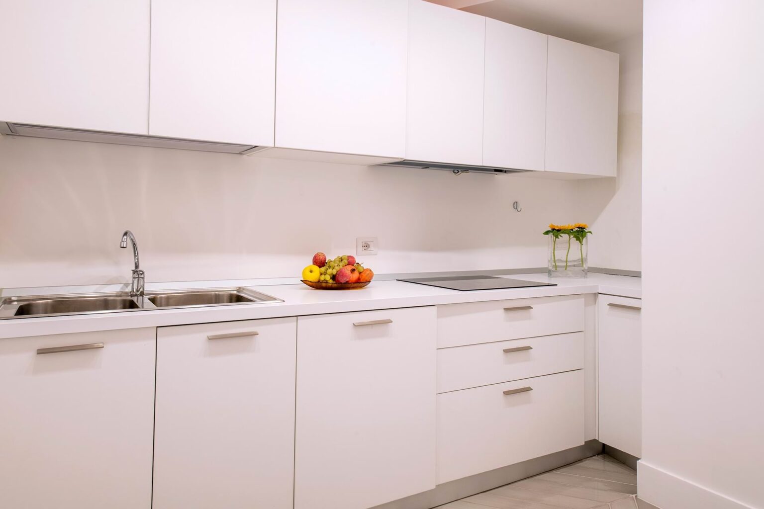 Modern white kitchen featuring a double sink, sleek cabinetry, and a minimalist design. A bowl of assorted fruits and a vase with flowers add a touch of color to the space. | Only on SopranoVillas
