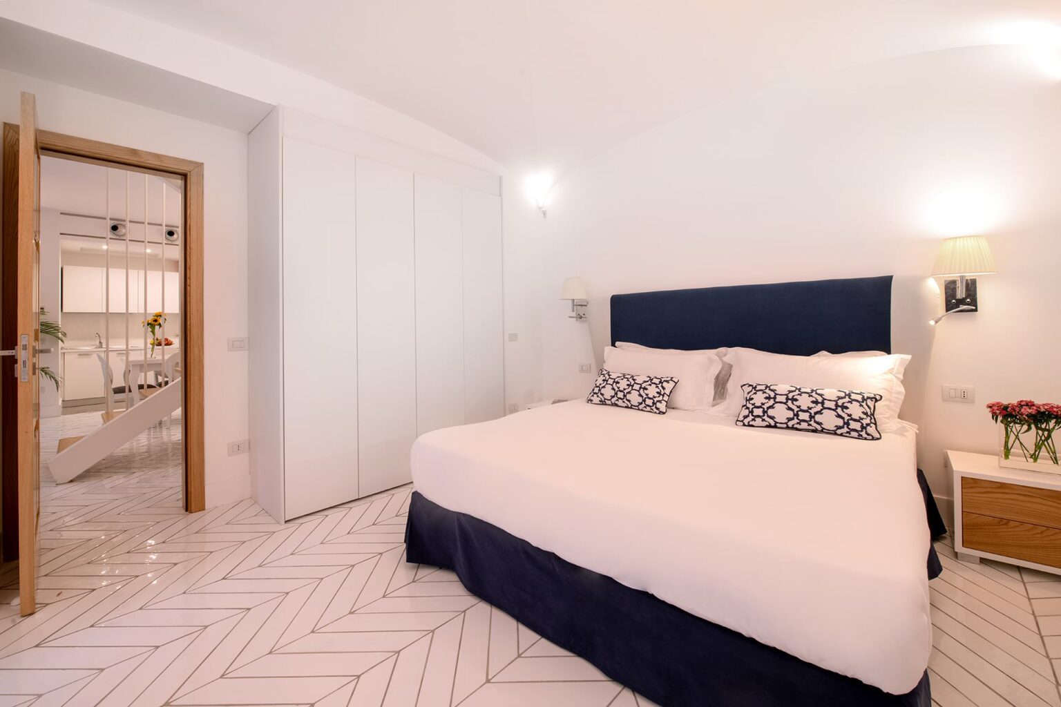 Modern bedroom featuring a comfortable queen-sized bed with decorative pillows, stylish nightstand, and a spacious wardrobe, all set in a bright, airy space with elegant chevron-patterned flooring. The open doorway leads to a well-lit living area. | Only on SopranoVillas