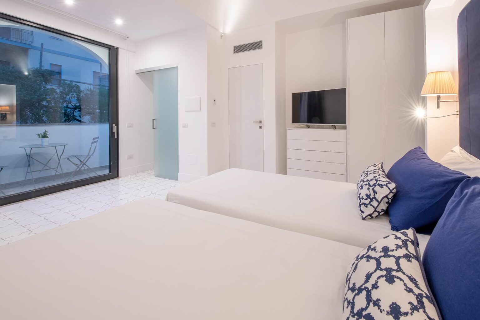 Modern hotel room featuring two beds, a flat-screen TV, and a sliding glass door leading to a balcony with outdoor furniture. The decor includes blue and white accents, bright lighting, and stylish furnishings, creating a comfortable and inviting atmosphere. | Only on SopranoVillas