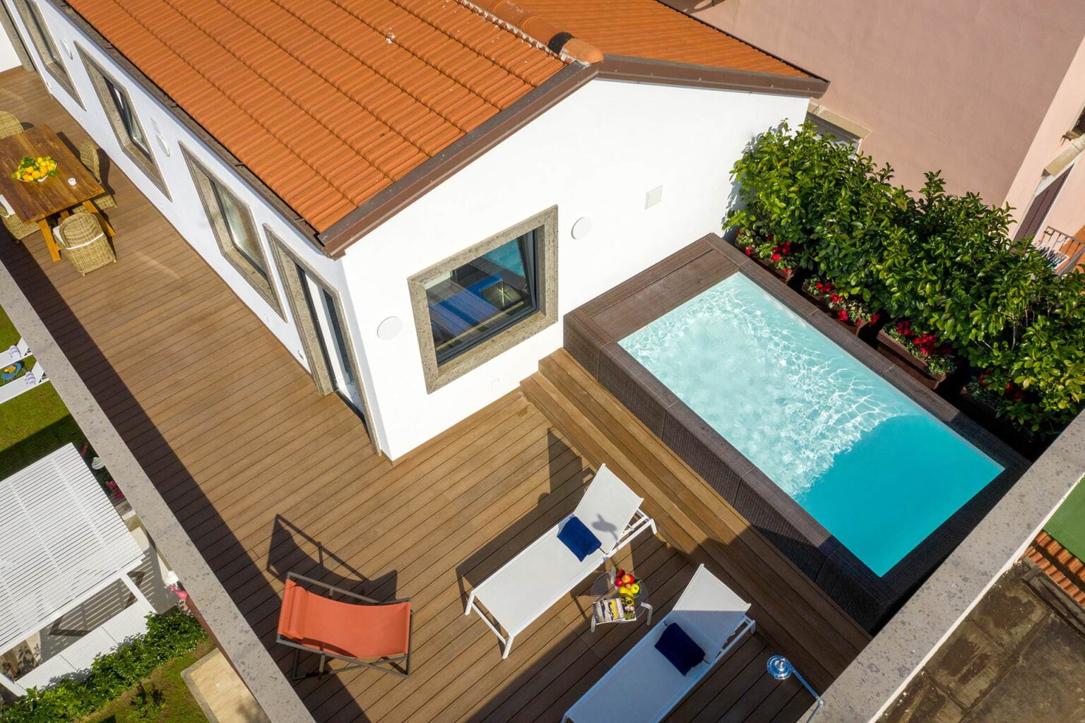 Aerial view of a modern house featuring a wooden deck with lounge chairs, a small swimming pool, and lush greenery surrounding the property. | Only on SopranoVillas