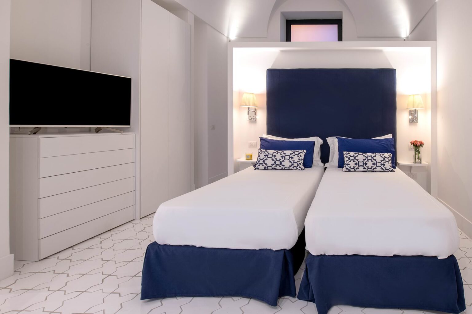 Modern hotel room featuring two single beds with blue and white bedding, minimalist white furniture, a flat-screen TV, and decorative accents. The stylish decor includes a geometric patterned floor and a small vase of flowers, creating a cozy atmosphere. Ideal for travelers seeking comfort and elegance. | Only on SopranoVillas