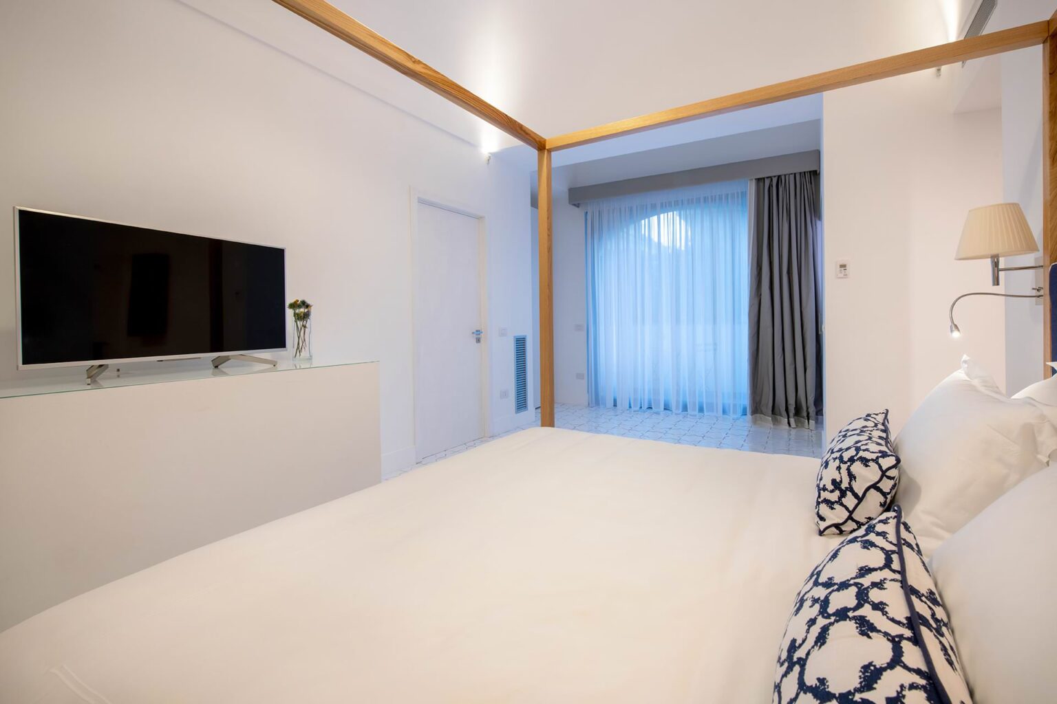 Modern hotel room featuring a cozy bed with decorative pillows, a flat-screen TV on a glass console, and large windows with sheer curtains allowing natural light. The minimalist decor and neutral colors create a serene atmosphere. | Only on SopranoVillas