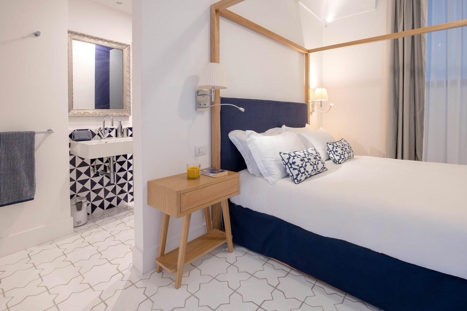 Cozy hotel room featuring a comfortable bed with decorative pillows, a bedside table, and a view into a stylish bathroom with patterned tiles. Bright and inviting atmosphere with natural light. | Only on SopranoVillas