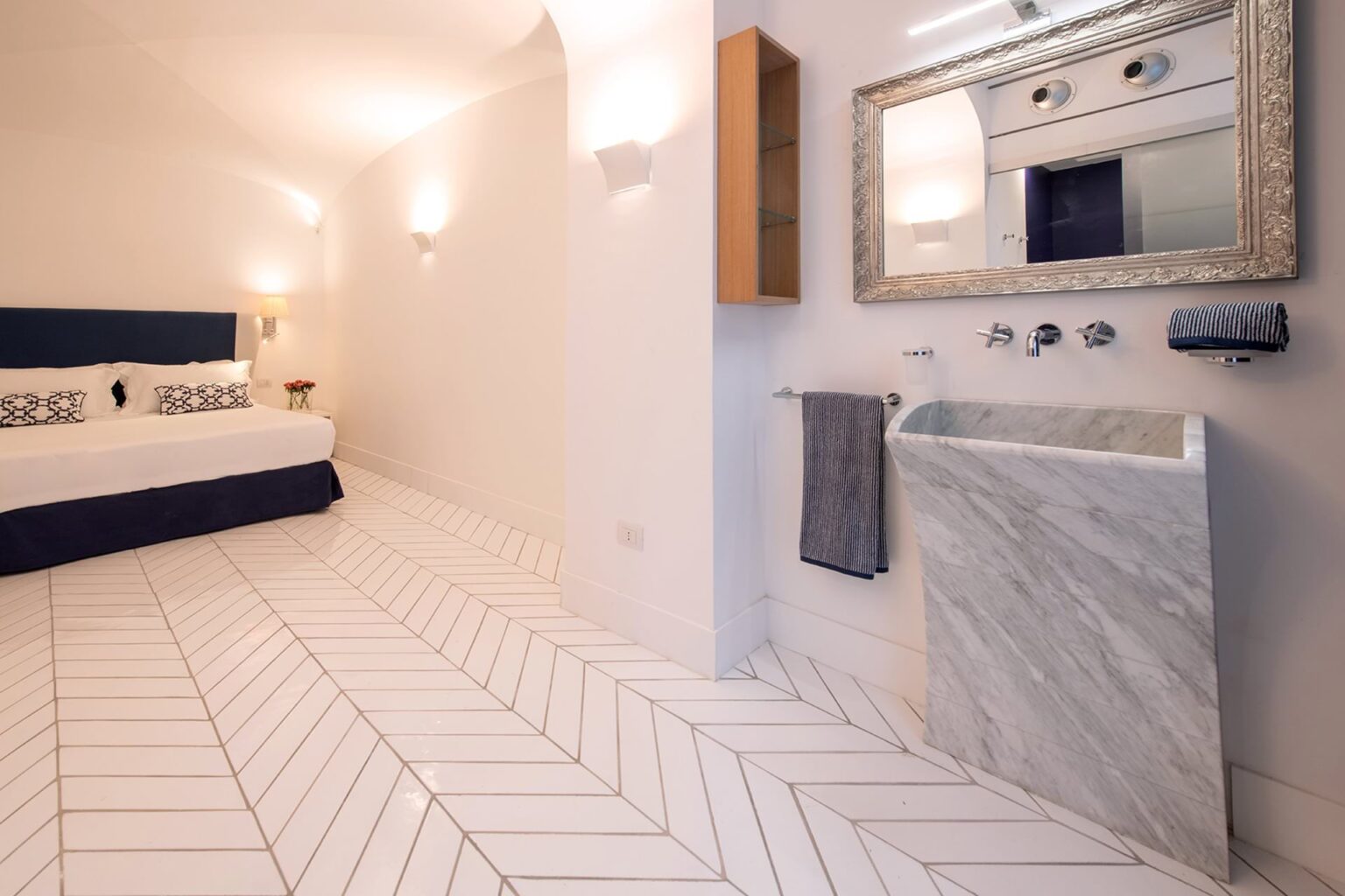 Modern hotel room featuring a double bed with decorative pillows, elegant marble sink, and stylish herringbone patterned floor tiles. The room is illuminated with soft lighting, enhancing its contemporary design. | Only on SopranoVillas
