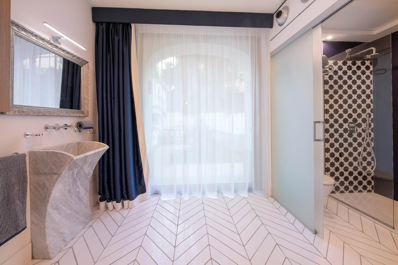 Modern bathroom interior featuring a stylish marble sink, elegant shower with glass door, and large windows with sheer curtains, complemented by herringbone patterned flooring and dark blue drapes. | Only on SopranoVillas