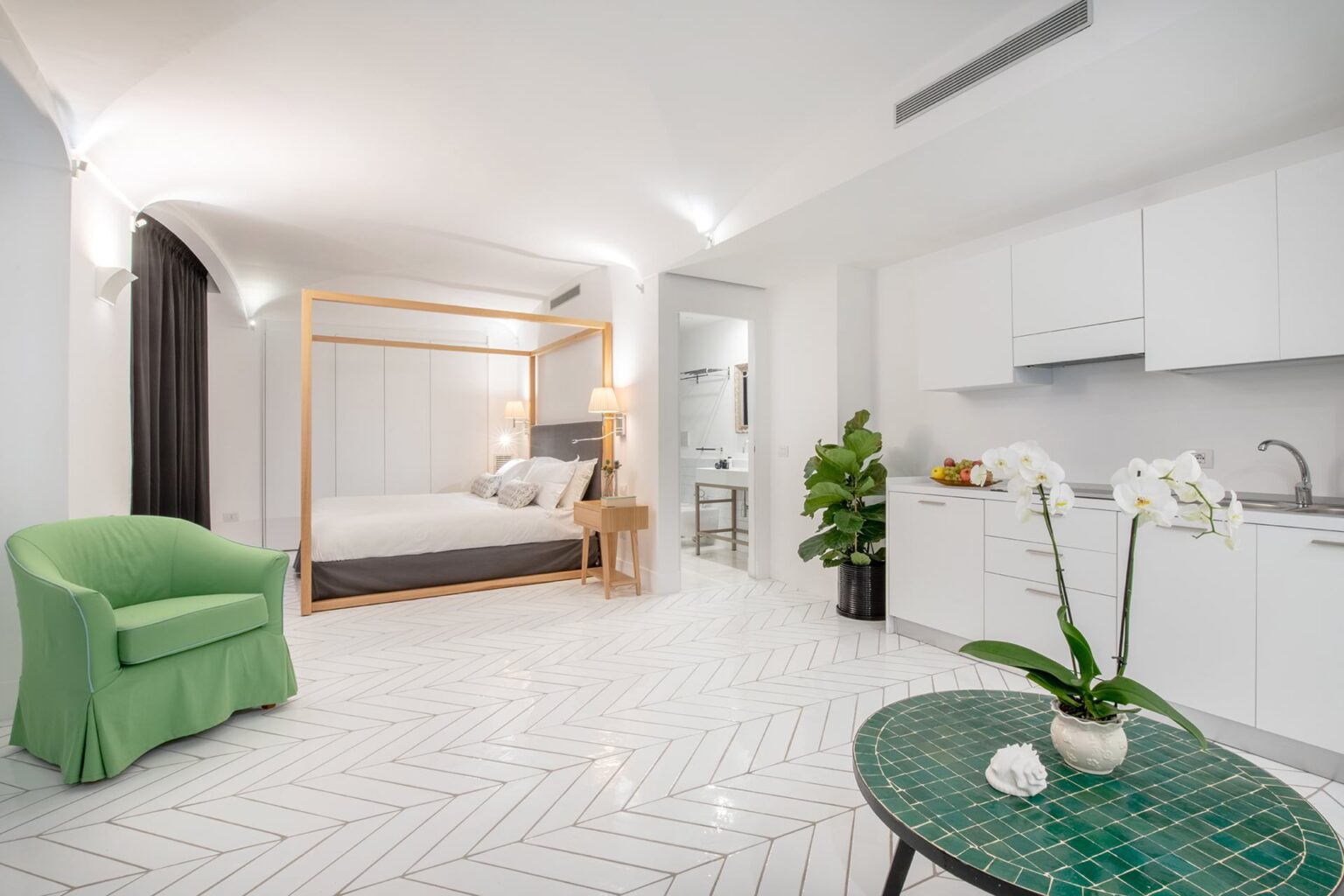 Modern studio apartment featuring a four-poster bed, green accent chair, and a kitchenette. The space is designed with white herringbone flooring, soft lighting, and decorative plants, creating a bright and inviting atmosphere. Ideal for travelers seeking comfort and style. | Only on SopranoVillas