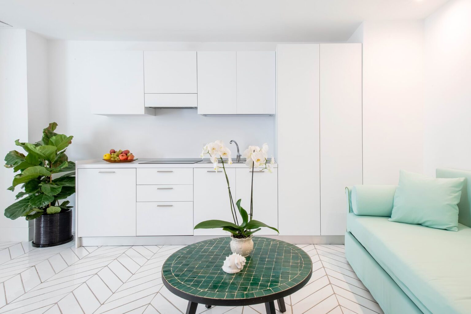 Modern kitchen and living area featuring white cabinetry, a green accent sofa, and a round coffee table with decorative plants. Bright, minimalist design with a geometric floor pattern. | Only on SopranoVillas