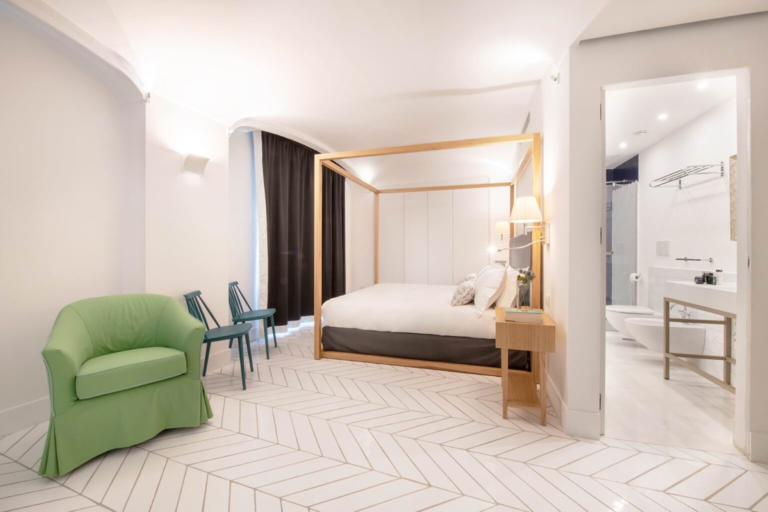 Modern hotel room featuring a four-poster bed, stylish green armchair, two blue chairs, and an en-suite bathroom with contemporary fixtures. The room has a light color scheme with herringbone flooring and ample natural light. | Only on SopranoVillas