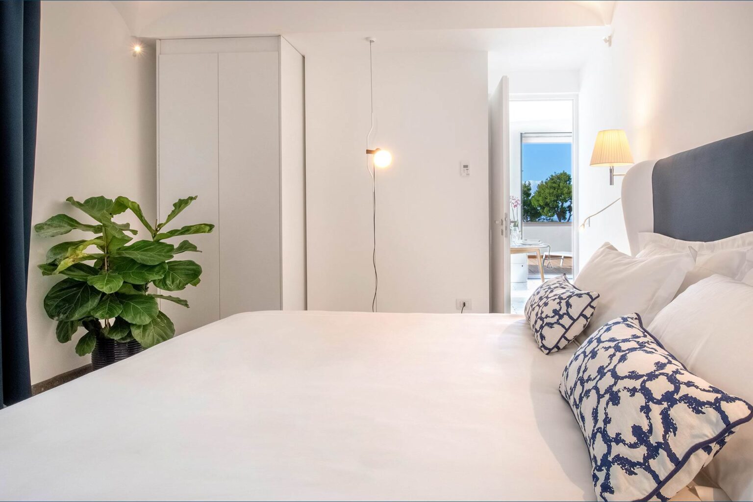 Modern bedroom interior featuring a spacious bed with decorative pillows, a stylish floor lamp, and a large window that offers natural light and a view of greenery. A potted plant adds a touch of nature to the decor. | Only on SopranoVillas