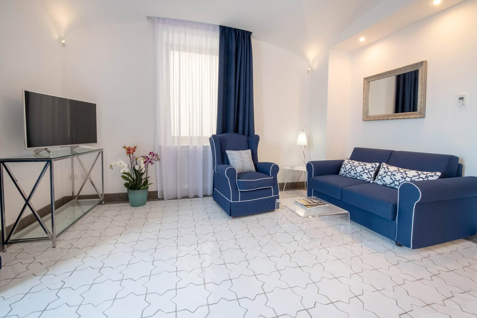 Modern living room featuring a blue sofa with decorative cushions, a glass TV stand, and a potted plant. Natural light streams through sheer curtains, highlighting the elegant tile flooring and stylish decor. | Only on SopranoVillas