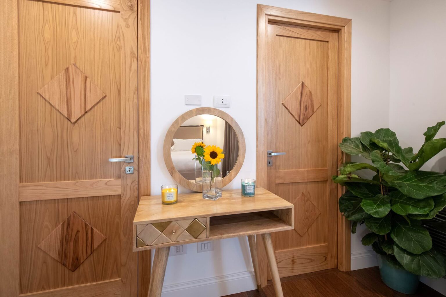 Modern entryway featuring a wooden console table with a round mirror, a vase of sunflowers, and decorative candles, flanked by two wooden doors with geometric designs and a green potted plant. | Only on SopranoVillas