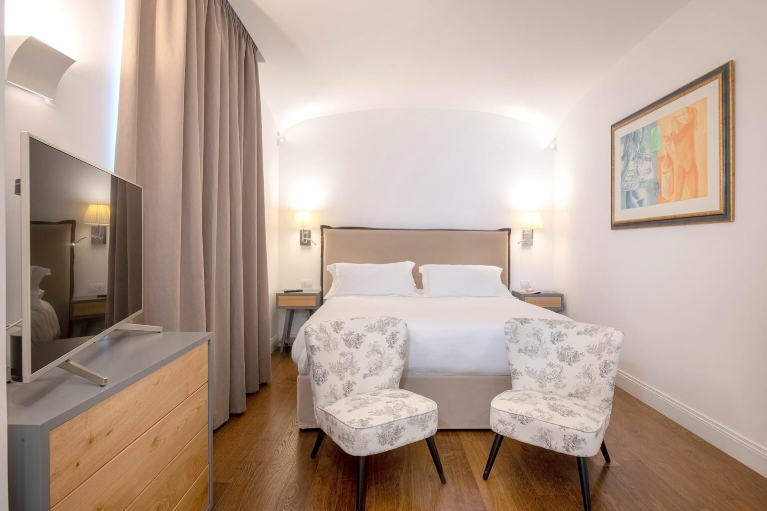 Modern hotel room featuring a comfortable queen-sized bed, stylish armchairs, a flat-screen TV, and elegant decor, ideal for relaxation and comfort. | Only on SopranoVillas
