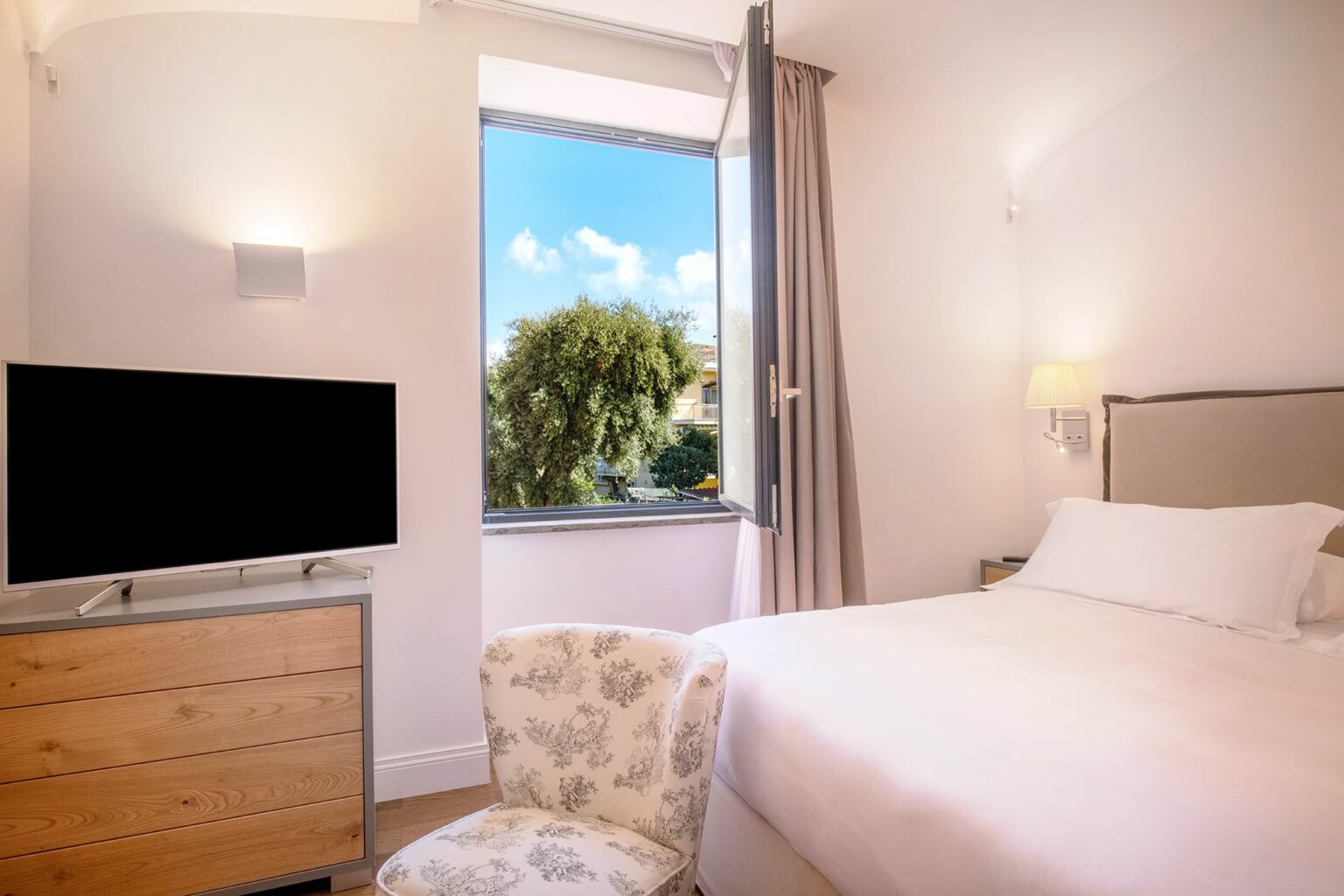 Bright and airy hotel room featuring a comfortable bed, a flat-screen TV, and a window with a view of greenery. The room is adorned with soft lighting and modern furnishings, creating a relaxing atmosphere. | Only on SopranoVillas